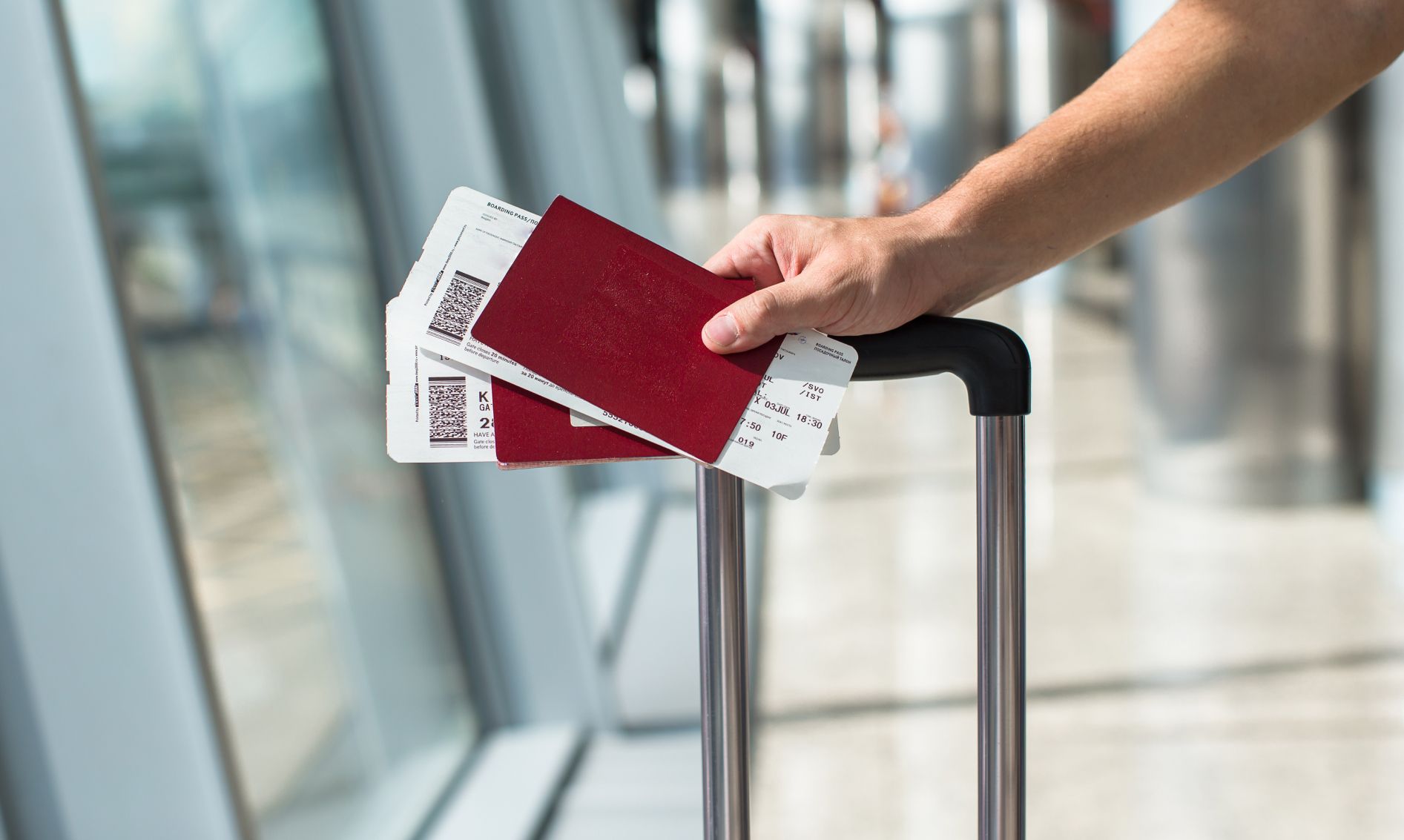 Close-up of a person holding two red passports and airline boarding passes while resting their hand on a luggage handle in a modern airport terminal.